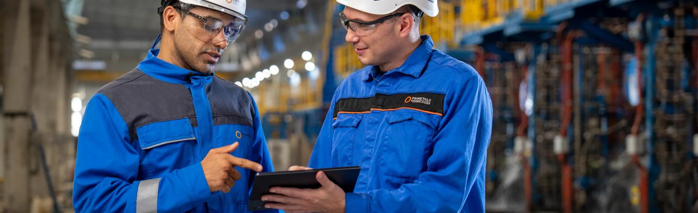 Two plant workers wearing safety gear review information together on a tablet inside an industrial facility
