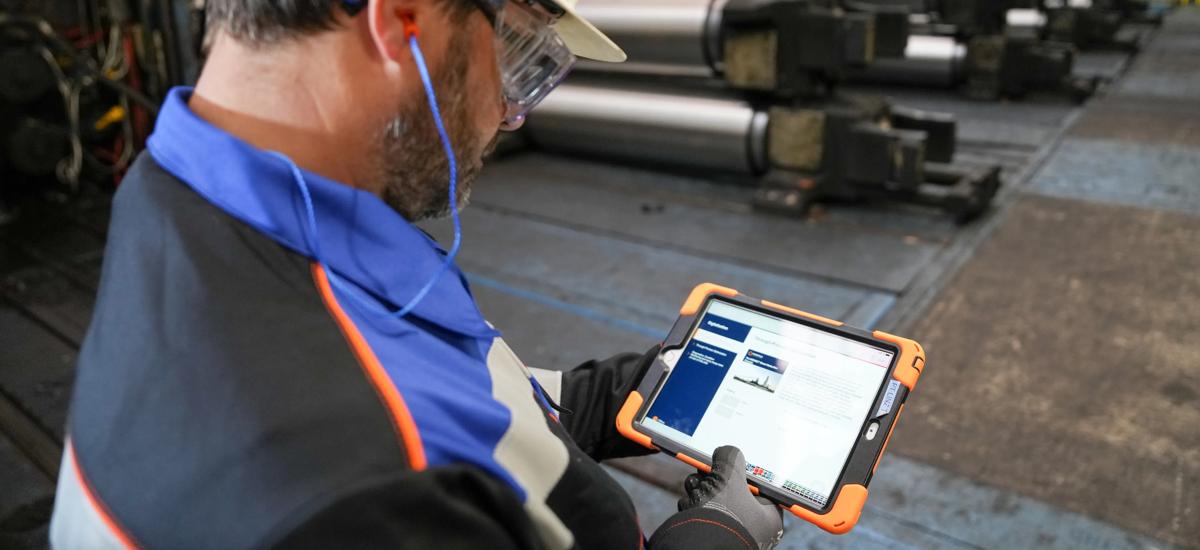 A service engineer inspects a rolling mill while reviewing data on a tablet, using digital tools to assess mill performance and support efficient maintenance and troubleshooting.