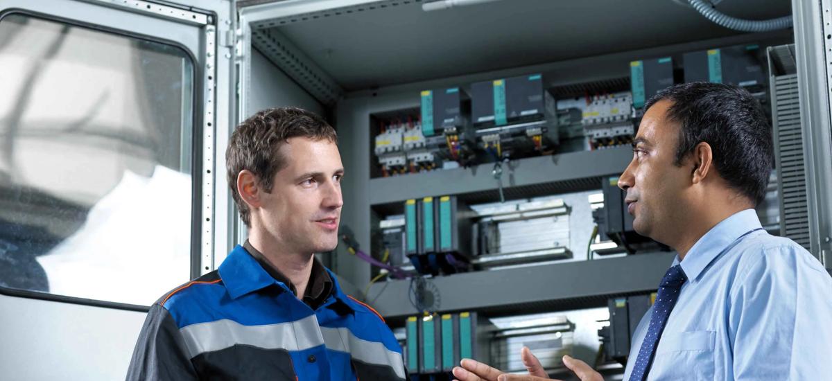 Service engineer explains the solution for a detected fault in front of the electrical PLC cubicle, ensuring reliable troubleshooting and plant operation.