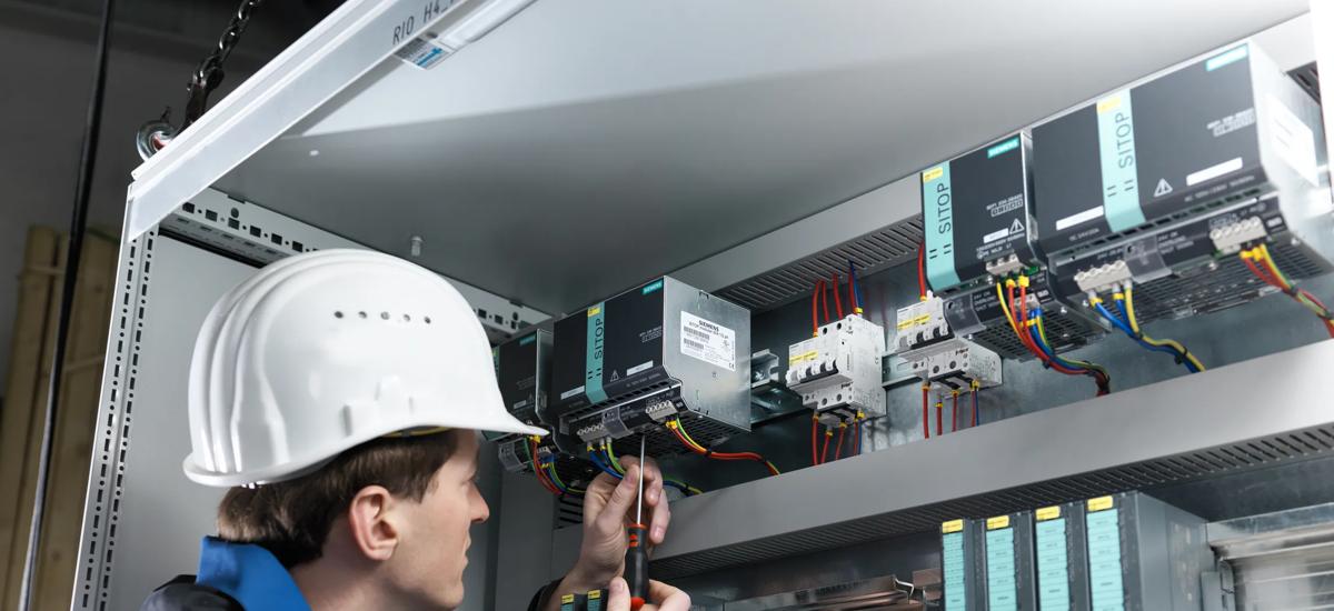 A service engineer troubleshoots inside an electrical PLC cubicle, inspecting components and connections to diagnose and resolve control system issues.